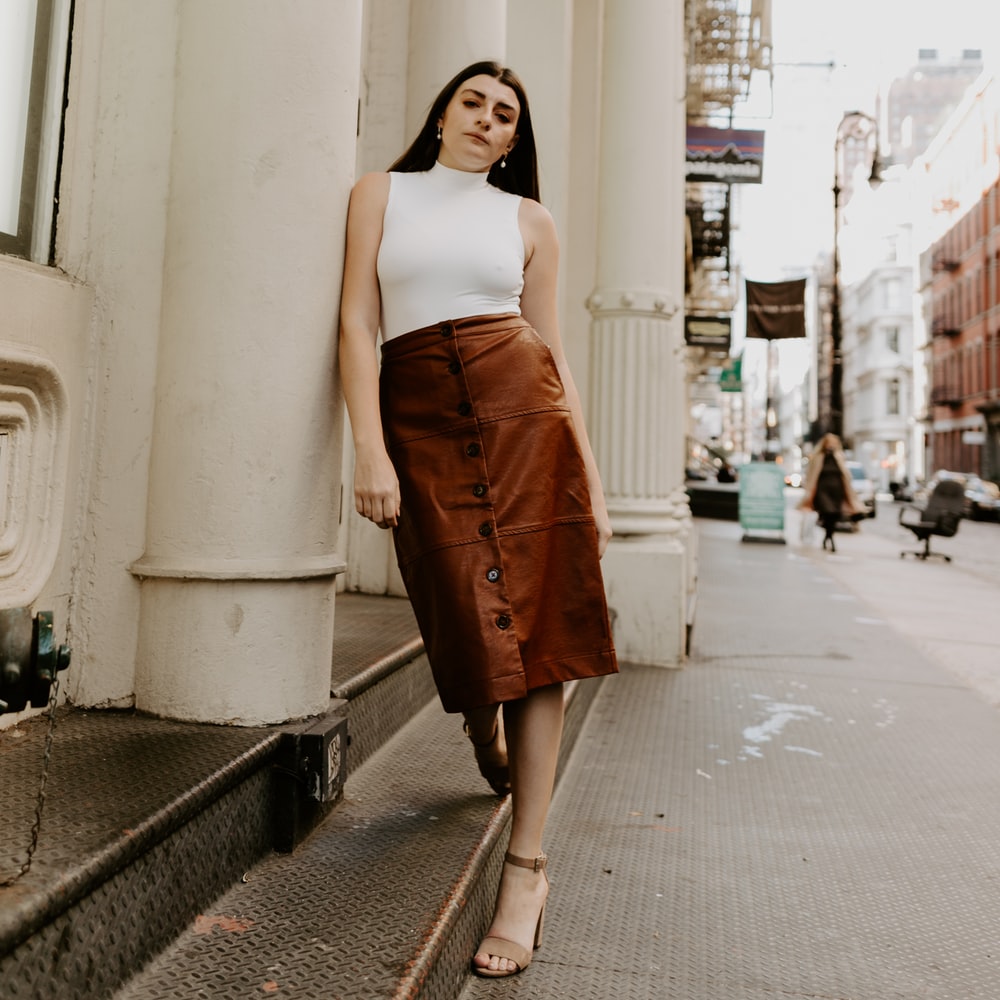 Woman In White Tank Top And Brown Skirt Standing On Sidewalk During Daytime