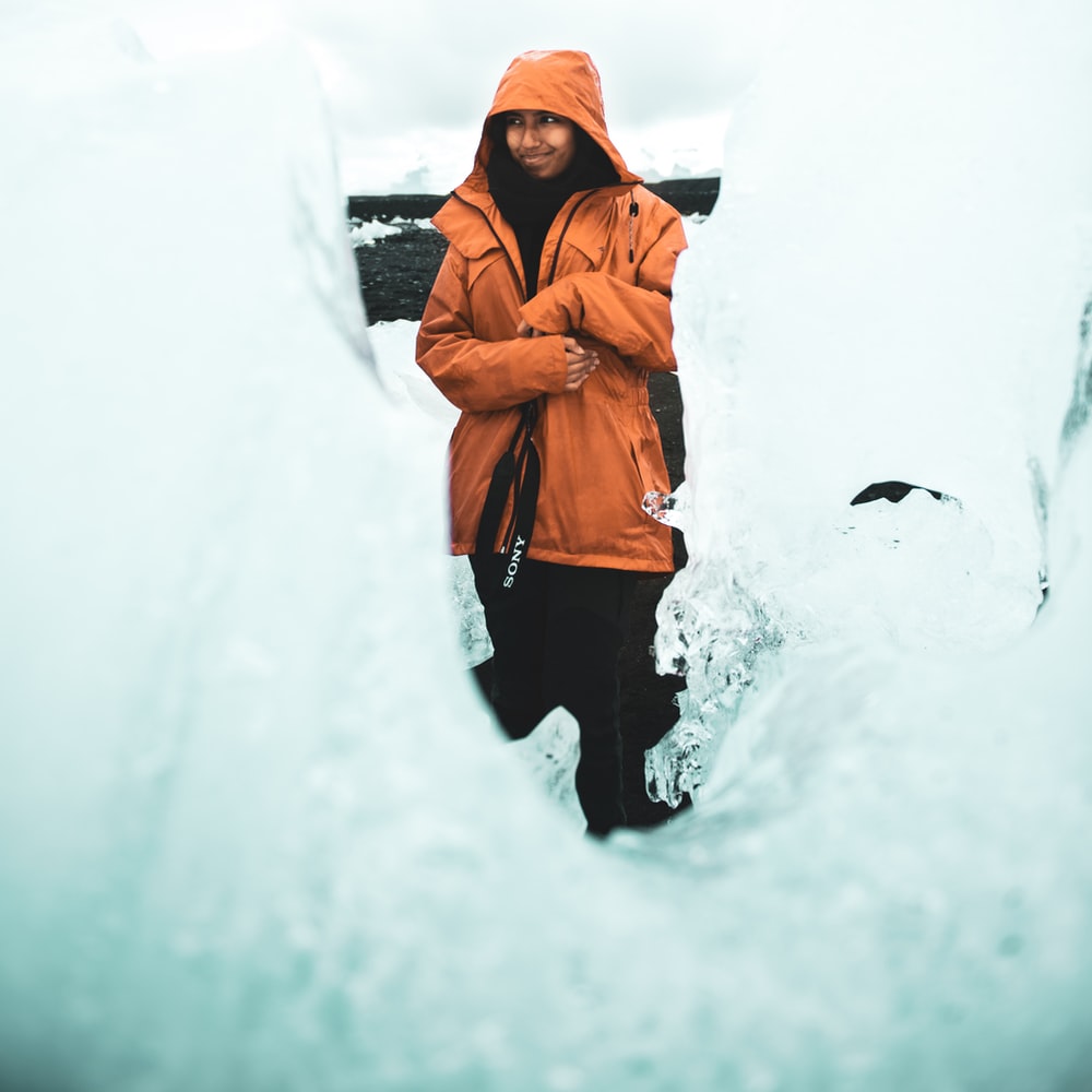 Woman In Orange Hoodie Standing On Snow Covered Ground During Daytime
