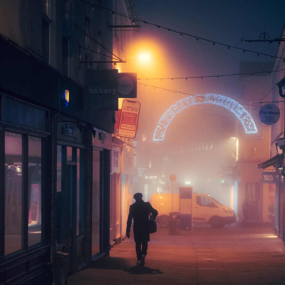 Man In Black Jacket Walking On Sidewalk During Night Time