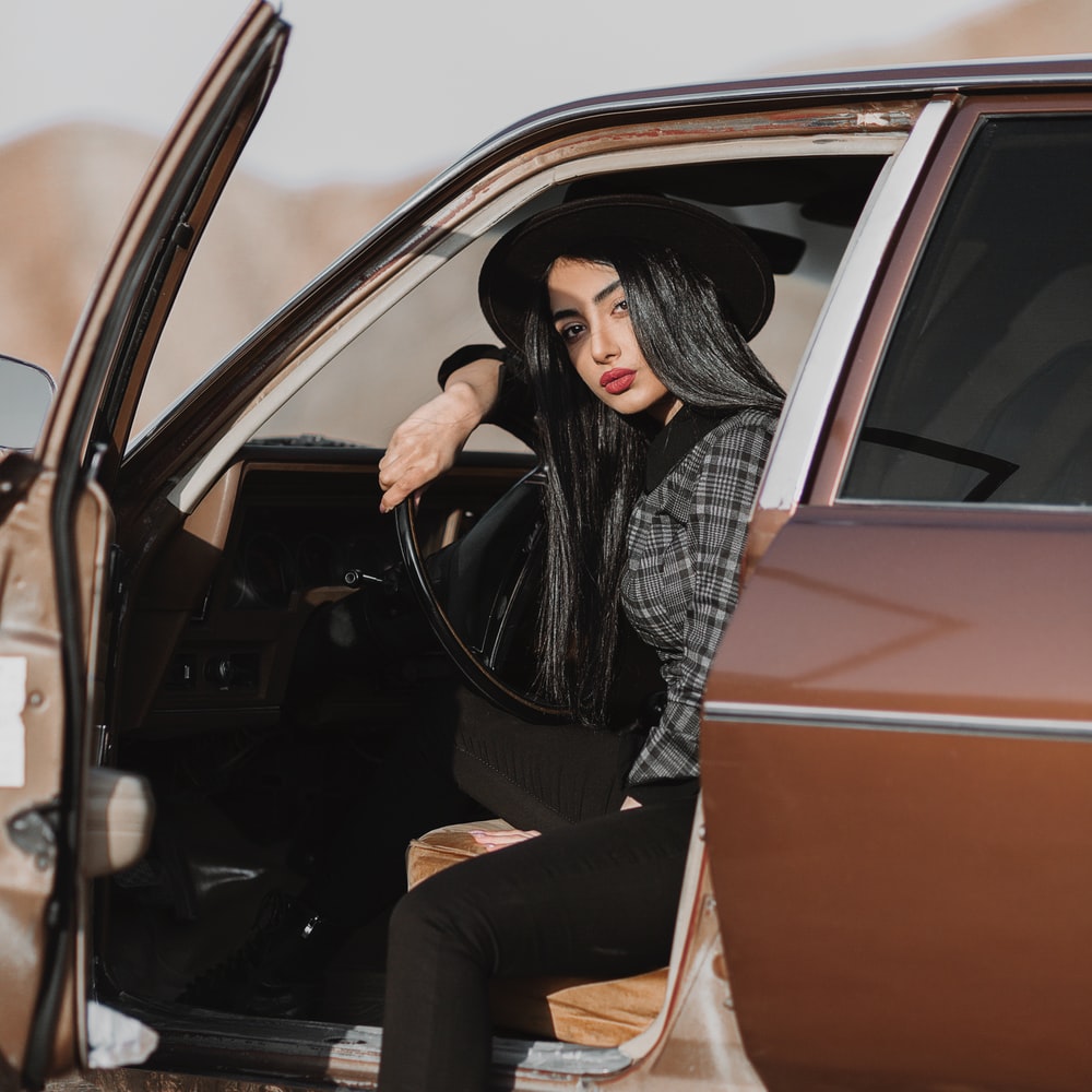 Woman In Black Leather Jacket And Black Pants Sitting On Car Door During Daytime