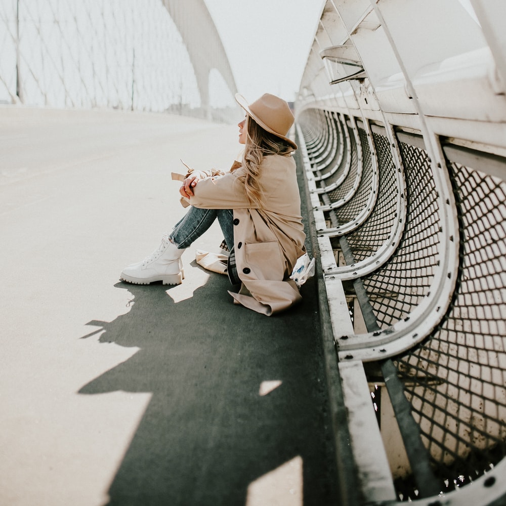 Woman In Brown Leather Jacket And Blue Denim Jeans Sitting On White Metal Fence During Daytime