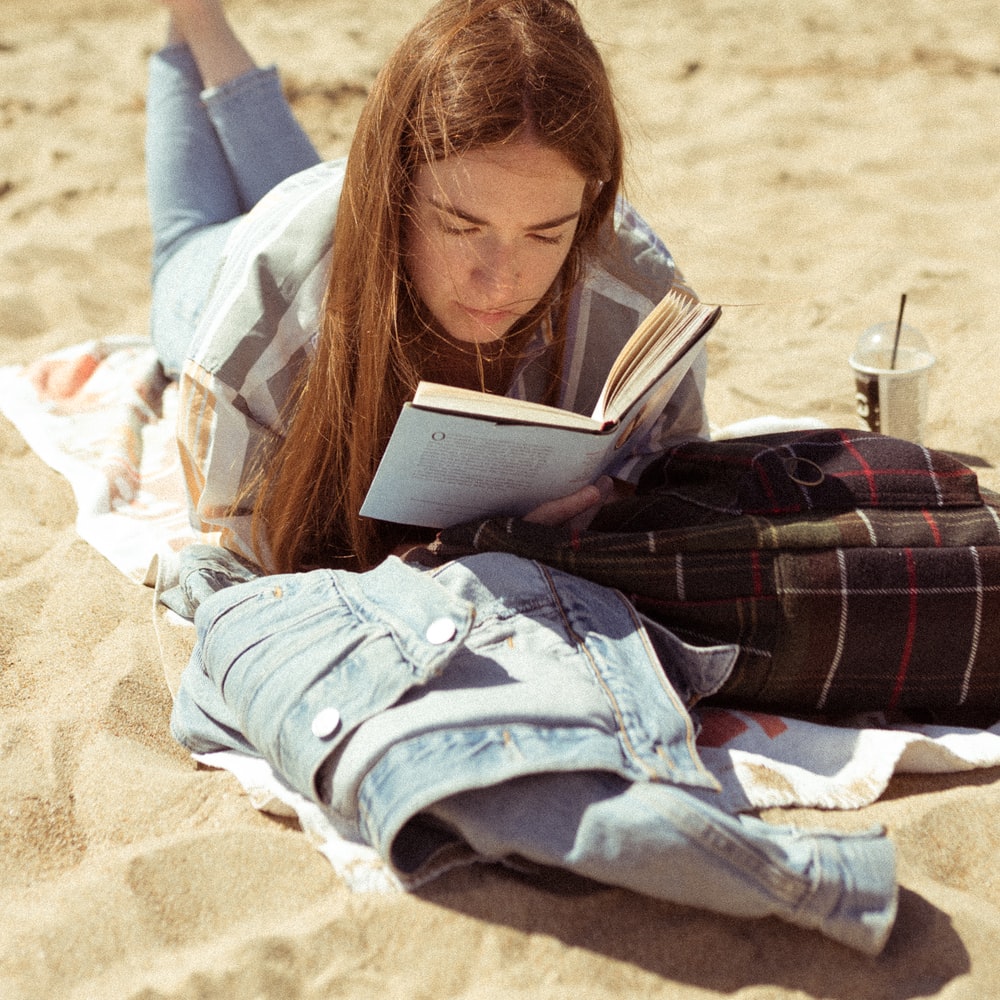Woman In White Long Sleeve Shirt Reading Book On Beach During Daytime