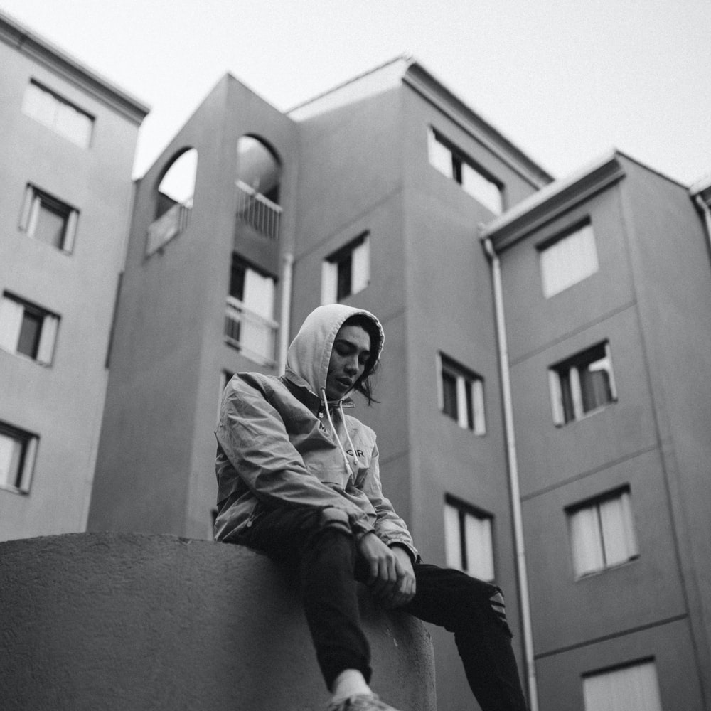 Grayscale Photo Of Man In Jacket And Pants Sitting On Concrete Wall