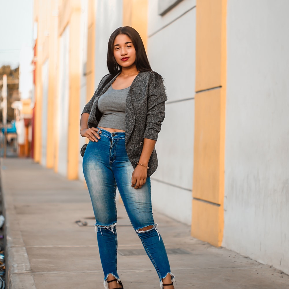 Woman In Gray Long Sleeve Shirt And Blue Denim Jeans Standing On Sidewalk During Daytime raster image